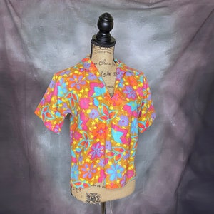 May include: A short-sleeved, collared shirt with a vibrant floral print in shades of orange, pink, blue, and yellow. The shirt has a V-neck and a relaxed fit.