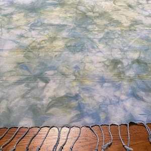 May include: A rectangular, tie-dye scarf in shades of blue, green, and white. The fabric has a textured appearance with horizontal lines and a fringe of knotted tassels along the bottom edge. The scarf is displayed on a wooden surface.