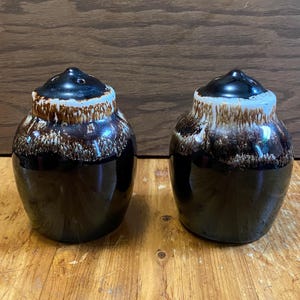 May include: A pair of brown and white salt and pepper shakers with a glazed finish. The shakers are round and have a small opening at the top for pouring.