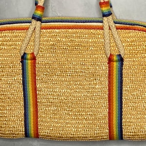 May include: A rectangular straw handbag with a woven texture. The bag features rainbow-colored stripes down the front and on the handles. The edges are trimmed with orange and blue stripes, and the handles are made of a thick, off-white rope.