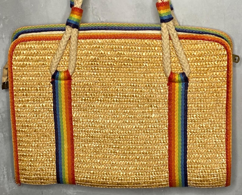 May include: A rectangular straw handbag with a woven texture. The bag features rainbow-colored stripes down the front and on the handles. The edges are trimmed with orange and blue stripes, and the handles are made of a thick, off-white rope.