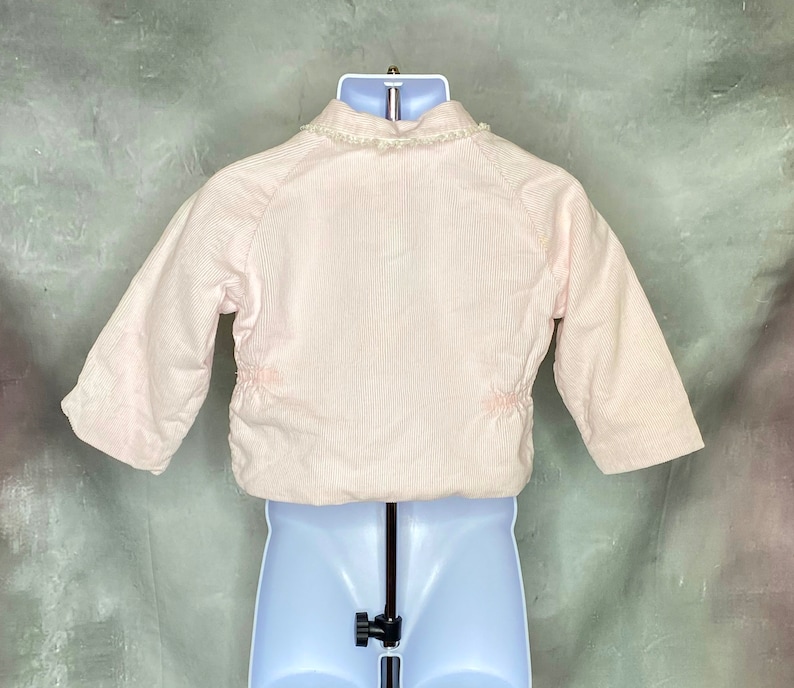 May include: A pink corduroy jacket with a white lace trim around the collar. The jacket has a zipper closure and long sleeves.