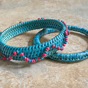 May include: Two turquoise woven bracelets with a beaded accent of pink and purple beads. The bracelets are circular and appear to be made of a woven material. The bracelets are on a neutral-toned surface.