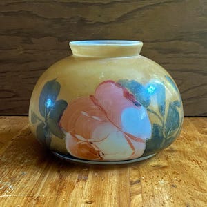 Peut inclure: Un vase rond en verre de couleur crème avec un motif floral peint à la main. Le vase présente une grande rose rose et orange avec des feuilles vertes.