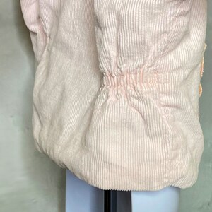 May include: A pink corduroy jacket with a cinched waist and a button closure. The jacket has a textured, ribbed fabric.