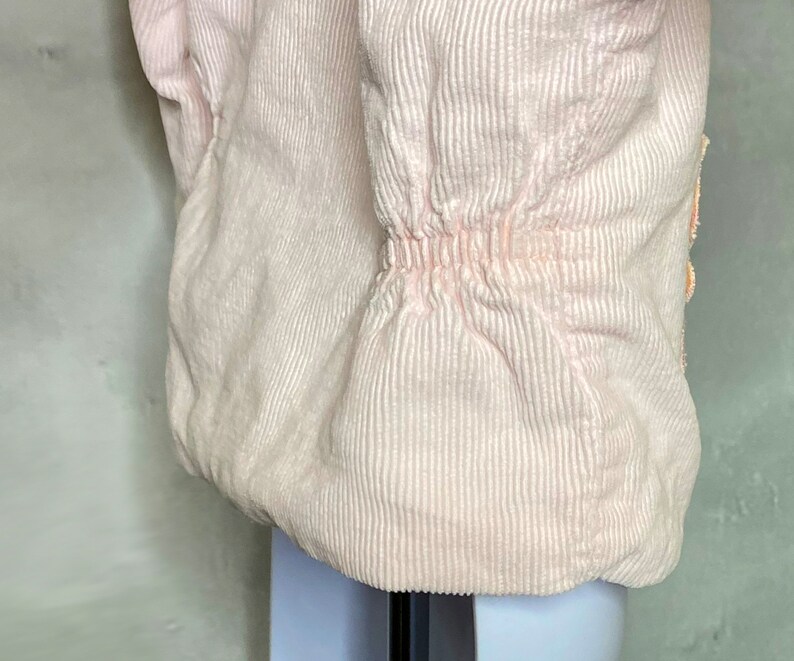 May include: A pink corduroy jacket with a cinched waist and a button closure. The jacket has a textured, ribbed fabric.