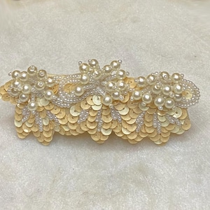 May include: A cream-colored hair clip with a design of overlapping sequins and pearl-like beads.