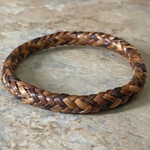 May include: Braided leather bracelet in shades of brown. The bracelet is circular and appears to be made of interwoven strips of leather. The colors range from light tan to dark brown, creating a textured appearance. A handcrafted accessory.