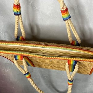May include: A woven straw tote bag with a rainbow trim and accents. The bag has a zipper closure and rope handles with rainbow details. The bag is open, revealing the interior lining.