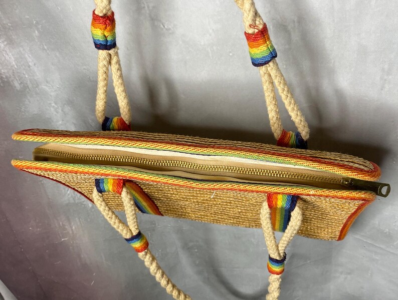 May include: A woven straw tote bag with a rainbow trim and accents. The bag has a zipper closure and rope handles with rainbow details. The bag is open, revealing the interior lining.