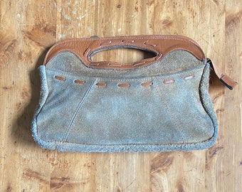 May include: A brown suede handbag with a brown leather flap and a top handle. The bag has a stitched design around the edges.