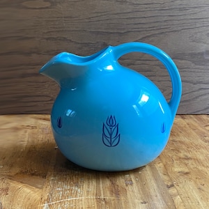 Könnte beinhalten: Eine hellblaue Keramikkanne mit Henkel. Die Kanne hat ein stilisiertes Blumenmuster in Blau auf der Vorderseite.
