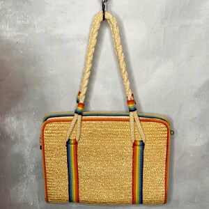 May include: A rectangular straw tote bag with a woven texture. The bag features a rainbow stripe design on the front and back, with matching rainbow accents on the braided handles. The bag is hanging from a black hook.