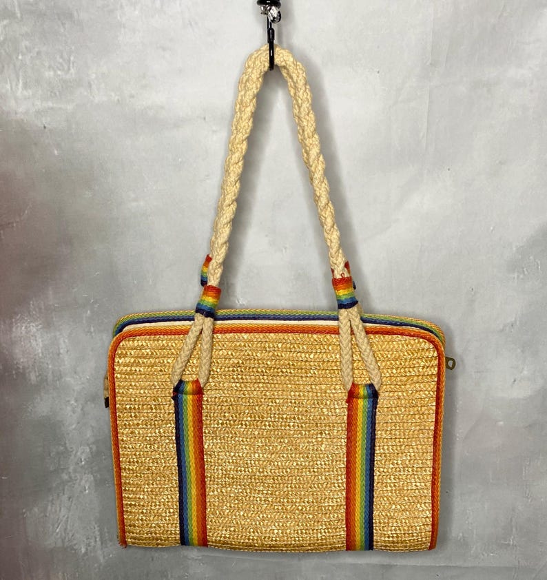 May include: A rectangular straw tote bag with a woven texture. The bag features a rainbow stripe design on the front and back, with matching rainbow accents on the braided handles. The bag is hanging from a black hook.