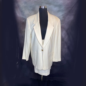 Può includere: Un tailleur bianco composto da giacca e gonna. La giacca ha una chiusura con un solo bottone e un revers a punta. La gonna è a trapezio e arriva al ginocchio.