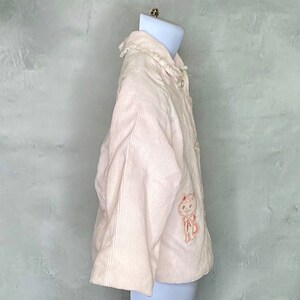 May include: A pink corduroy jacket with a white lace collar and a small embroidered animal on the front.