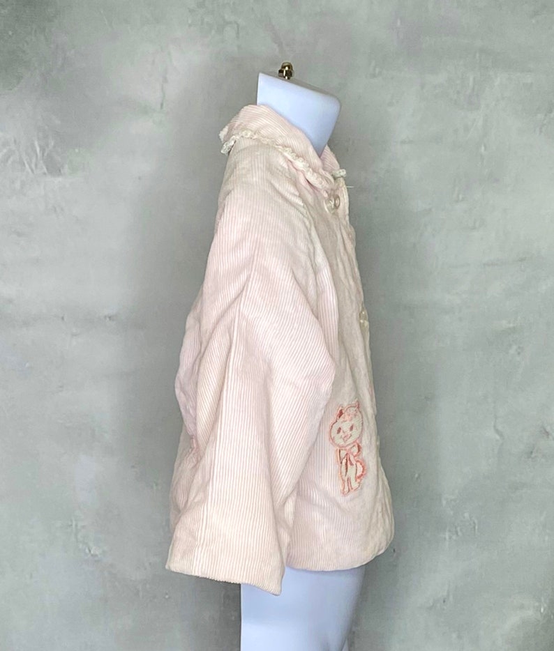 May include: A pink corduroy jacket with a white lace collar and a small embroidered animal on the front.