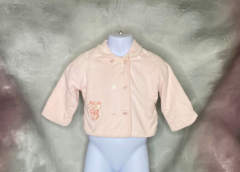 May include: A pink corduroy baby jacket with a white lace collar and a small embroidered cat on the front.