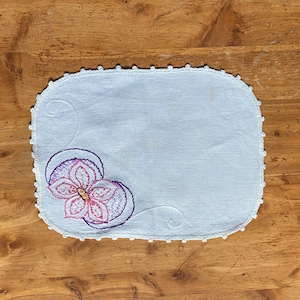 May include: White linen embroidered with a purple and pink flower with a yellow center. The linen has a white crocheted border.