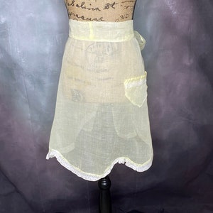 May include: A pale yellow apron with a white lace trim and a single pocket. The apron is tied at the waist and has a scalloped hem.