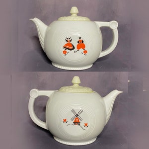 Puede incluir: Tetera de cerámica blanca con tapa y asa color crema. La tetera presenta un diseño circular con ilustraciones de una pareja con vestimenta tradicional holandesa y un molino de viento. Tiene un pico vertedor y un asa curva.
