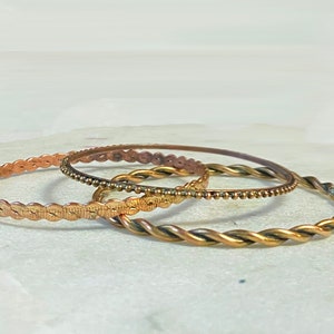 Pode incluir: Três pulseiras de bracelete douradas. As pulseiras estão empilhadas uma em cima da outra. A pulseira superior tem um design de contas, a pulseira do meio tem um design trançado e a pulseira inferior tem um design festoneado.