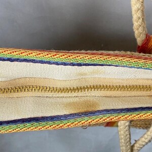 May include: A rainbow-striped tote bag with rope handles. The bag features a cream-colored interior with a gold zipper closure. The exterior has a woven design with rainbow stripes and a blue border.