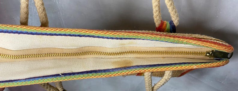 May include: A rainbow-striped tote bag with rope handles. The bag features a cream-colored interior with a gold zipper closure. The exterior has a woven design with rainbow stripes and a blue border.