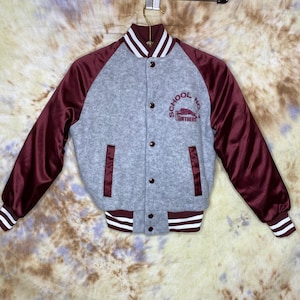 May include: A gray and maroon varsity jacket with a white stripe on the collar and cuffs. The jacket has a maroon satin lining and a maroon pocket on the left side. The back of the jacket has a maroon embroidered logo that says "School No. 50 Panthers".