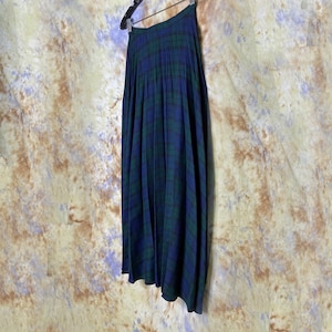 May include: A long, pleated skirt in a black and green tartan pattern. The skirt is hanging on a hanger and has a dark blue background.
