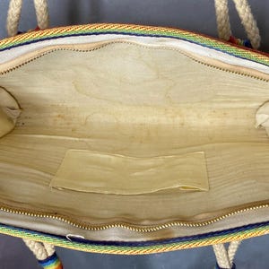 May include: Open view of a vintage handbag with a rainbow trim and rope handles. The interior is beige with a small pocket. The zipper is gold-colored. The bag is rectangular and appears to be made of fabric.