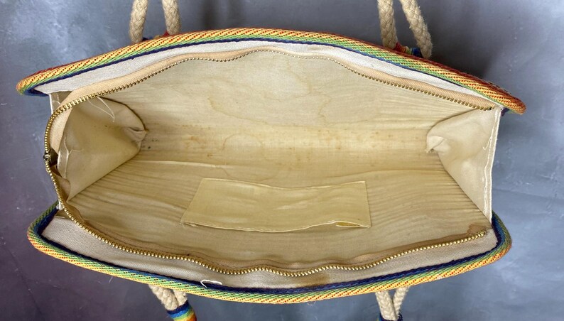 May include: Open view of a vintage handbag with a rainbow trim and rope handles. The interior is beige with a small pocket. The zipper is gold-colored. The bag is rectangular and appears to be made of fabric.