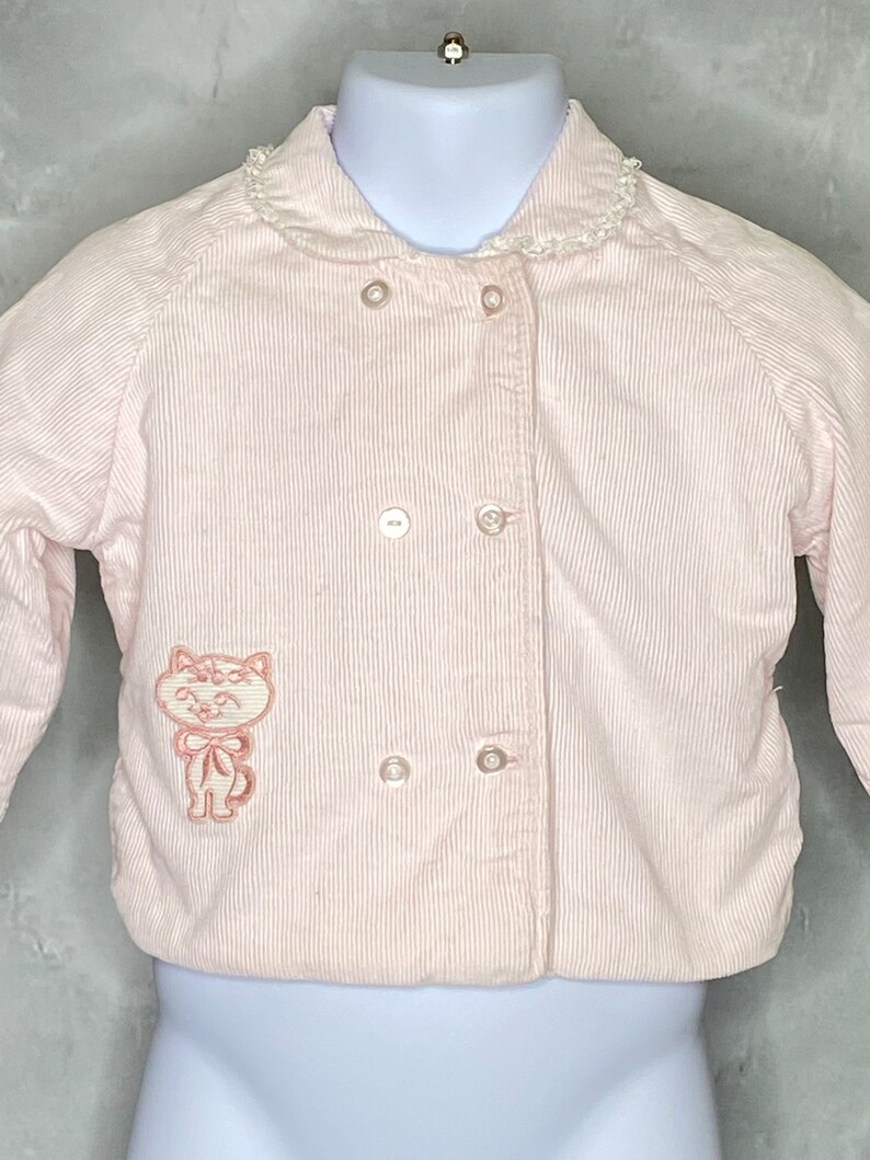 May include: A pink corduroy jacket with a white lace collar and a pink embroidered cat on the front.