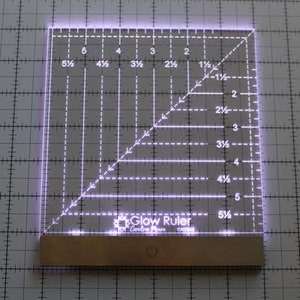 May include: A white acrylic illuminated ruler with a grid pattern and markings in inches and fractions of an inch. The ruler has a built-in LED light and a power button. The text "Glow Ruler" is printed on the ruler.