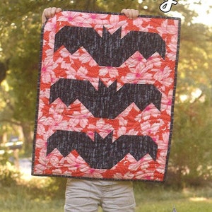 May include: A mini quilt with a floral pattern in red and pink, featuring three black bat silhouettes. The quilt is held up, with the text "Lil' Batty" and "Running Doe Quilts" visible. The background is blurred.