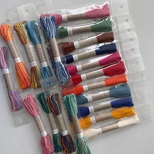 May include: A collection of colorful embroidery floss skeins in various shades, including pink, orange, blue, and green. Each skein is wrapped with a paper label and packaged in clear plastic. Ideal for embroidery and cross-stitch projects.