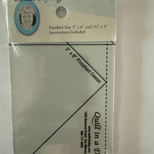 May include: A clear plastic quilting ruler for making small flying geese. The packaging reads "Quilt in a Day" and "Small Flying Geese Ruler." The ruler is marked with lines and angles for precise cutting. Finished size is 3" x 6" and 1 1/2" x 3".