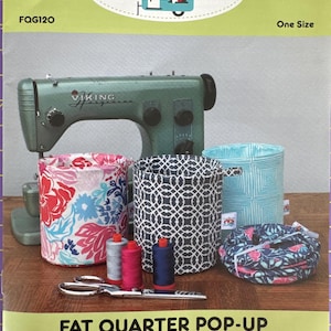 May include: Image of a sewing-themed craft project. The image features a green sewing machine, several fabric containers with floral and geometric patterns, spools of thread, and scissors. The text reads "FAT QUARTER POP-UP".