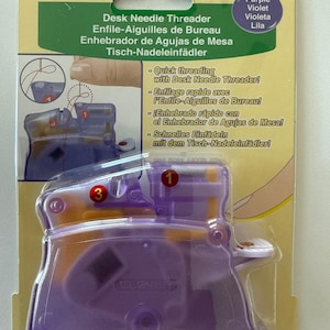 May include: A Clover brand Desk Needle Threader in purple and orange plastic, packaged on a yellow card. The packaging features the text Desk Needle Threader, 4071, and instructions with numbered diagrams for quick threading of sewing needles.