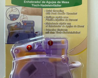 May include: A Clover brand Desk Needle Threader in purple and orange plastic, packaged on a yellow card. The packaging features the text Desk Needle Threader, 4071, and instructions with numbered diagrams for quick threading of sewing needles.