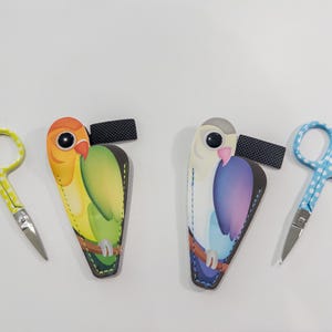 May include: Two pairs of nail clippers with colorful bird-shaped cases. One case is yellow, green, and orange with a parrot design. The other case is white, blue, and purple with a bird design. Both cases have a black strap.