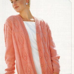 May include: A peach-colored knit cardigan featuring a V-neckline, long sleeves, and a button-front closure. The garment displays a textured diamond pattern with vertical cable knit detailing. The text INCLUDES LARGER SIZES is visible.