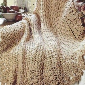 May include: A beige crochet blanket featuring a textured stitch pattern and a decorative scalloped border. The fabric has a consistent light tan color throughout, showcasing intricate yarn work with openwork details along the edges of the textile.