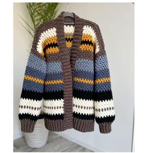 May include: A long-sleeve crochet cardigan featuring horizontal stripes in brown, cream, mustard yellow, light blue, and black. The garment has a solid brown ribbed collar and cuffs, an open front design, and a textured stitch pattern throughout.