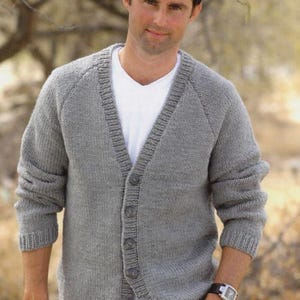 May include: A grey long-sleeve knit cardigan featuring a V-neckline, a button-down front with five visible buttons, and ribbed cuffs and hem. The garment is worn over a plain white V-neck t-shirt, showcasing a classic, textured knit construction.
