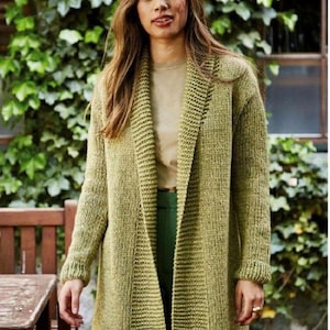 May include: A long-sleeved, open-front cardigan in a textured olive green knit. The garment features a wide, ribbed shawl collar, ribbed cuffs, and a straight hemline that falls below the hips, worn over a plain tan top and dark green trousers.