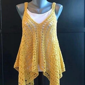 May include: A sleeveless yellow crochet top featuring an openwork pattern with a V-neckline and an asymmetrical hemline. The garment is layered over a plain white tank top, showcasing a loose, flowing silhouette with a textured, knotted stitch design.