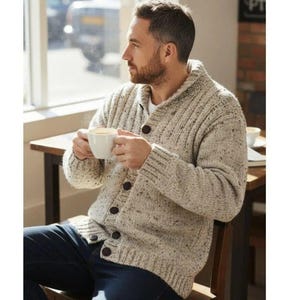 May include: A beige, speckled, chunky knit cardigan featuring a shawl collar, long sleeves, and a front button closure with five dark brown buttons. The knit fabric has a textured appearance with small dark flecks throughout the material.