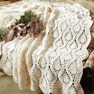 May include: A cream-colored crochet blanket featuring an intricate pineapple stitch pattern with scalloped edges. A small bouquet of dried flowers tied with a light pink ribbon rests on the folded fabric, adding a soft accent to the textile.