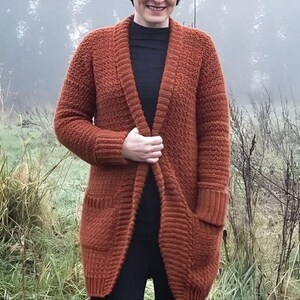 May include: A rust-colored, long-sleeved, open-front cardigan featuring a textured knit pattern, a ribbed collar, ribbed cuffs, and two large patch pockets on the front. The garment is worn over a plain black shirt and black pants.